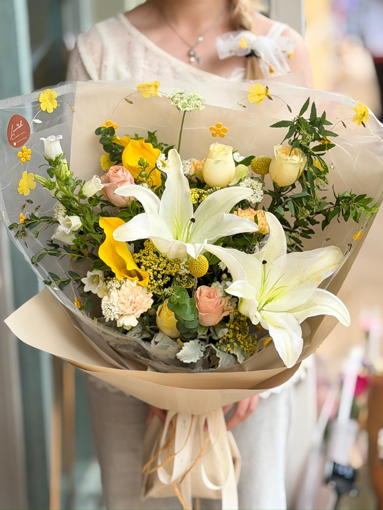 Honeydew Lily 百合花束近拍,展示了盛開的白百合、黃色海芋、奶油玫瑰與黃色跳舞蘭,採用現代奶油風半透明印花紙包裹。