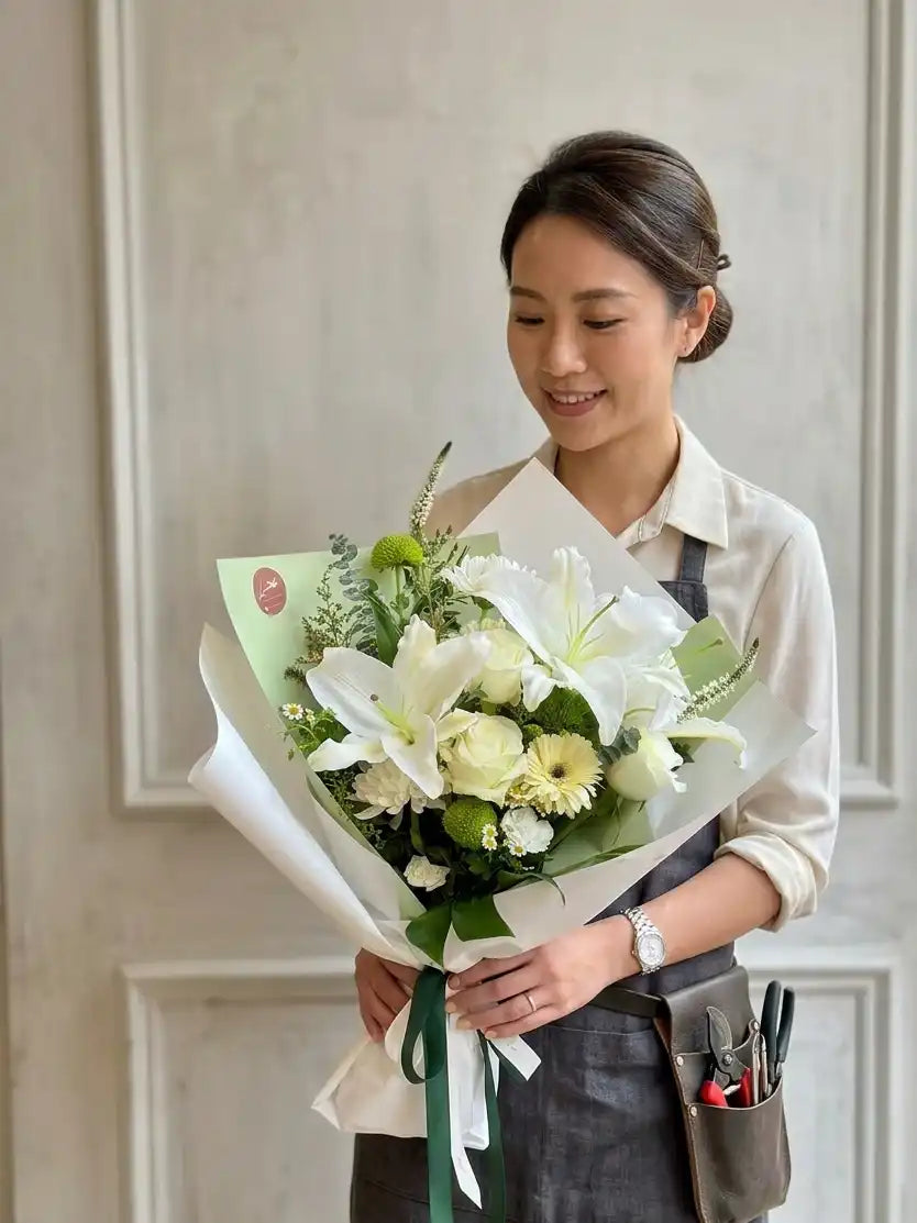 A professional florist with an apron presents a stunning, vibrant, hand-tied white and green lily bouquet, complete with roses and various textured foliage. Perfect floral arrangement for a clean, botanical aesthetic.