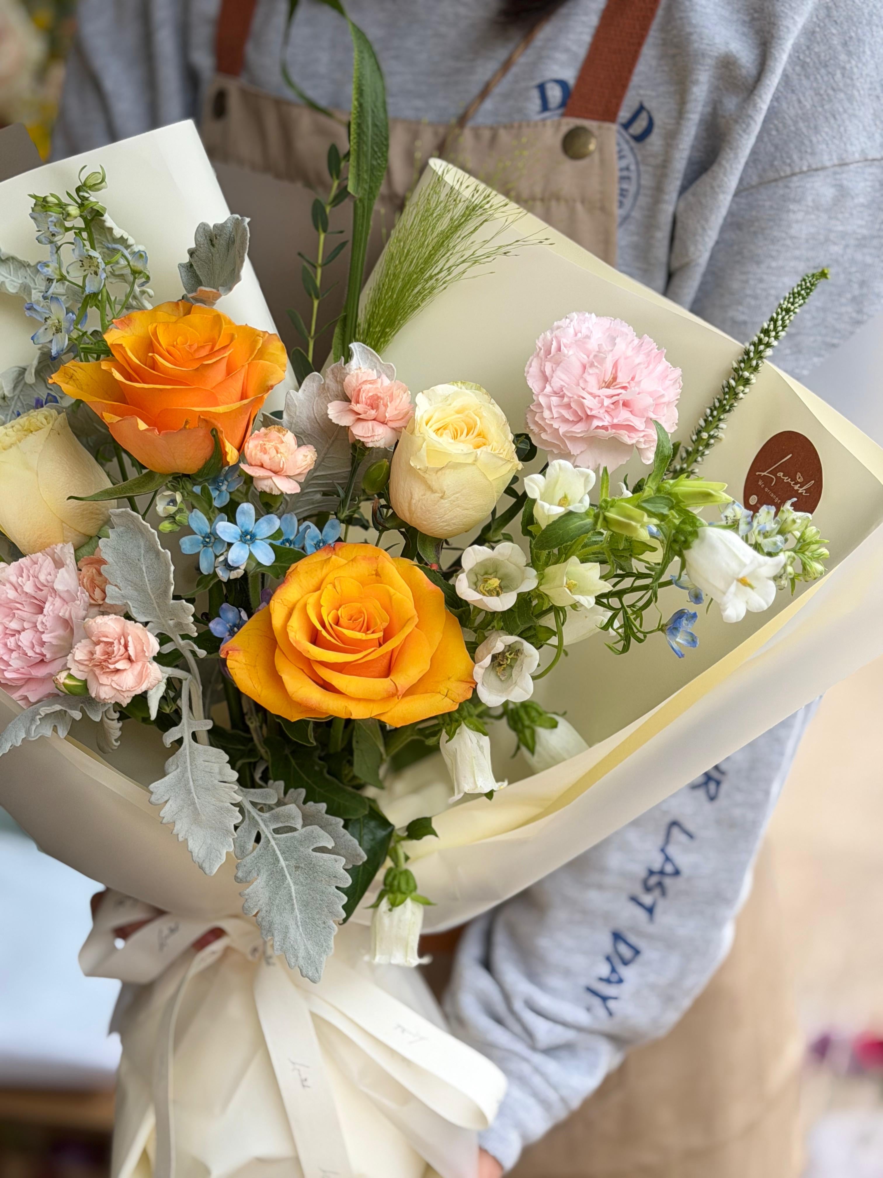 A close-up of the "Amber Muse" bouquet featuring vibrant orange roses, cream roses, and soft pink carnations. The arrangement includes delicate blue star-flowers and silvery dusty miller leaves, wrapped in elegant frosted cream and taupe paper with a translucent ribbon.