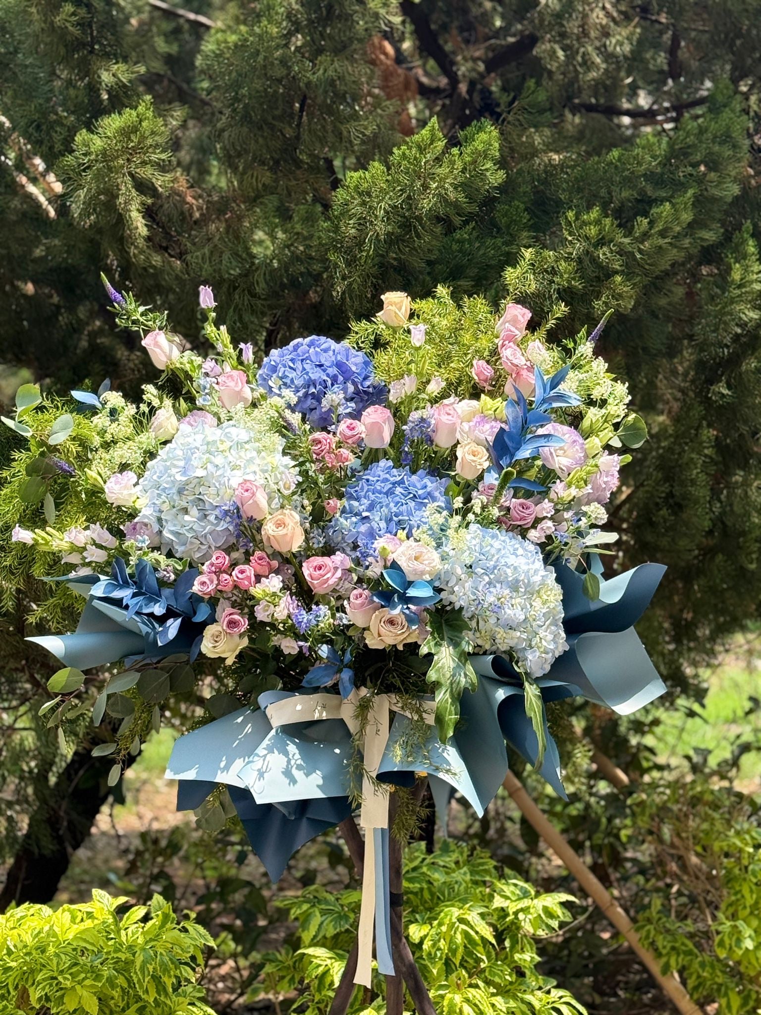 由Lavish Florist 設計 - Laurel Fleur Meadow 法式田園風開張花籃全貌展示,精選高貴大氣的深藍與淺藍色繡球花,交錯淡粉色玫瑰與白色風鈴草,搭配莫蘭迪藍色質感包裝紙,是一款專為 Cafe 及服飾店開幕設計的開張花籃。