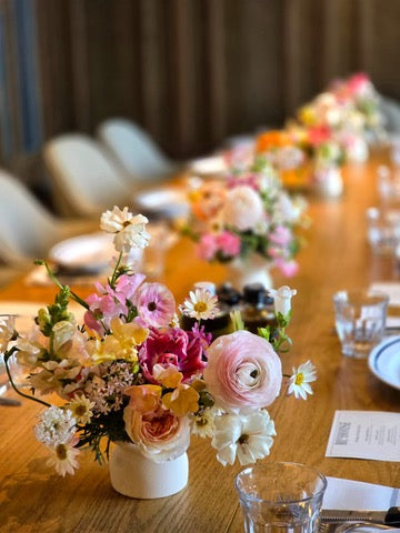 Indoor wedding tablescape by Lavish Florist featuring a long wooden dining table decorated with a row of vibrant floral centerpieces including pink ranunculus, pastel roses, and daisies in white ceramic vases, paired with elegant glassware and menus.