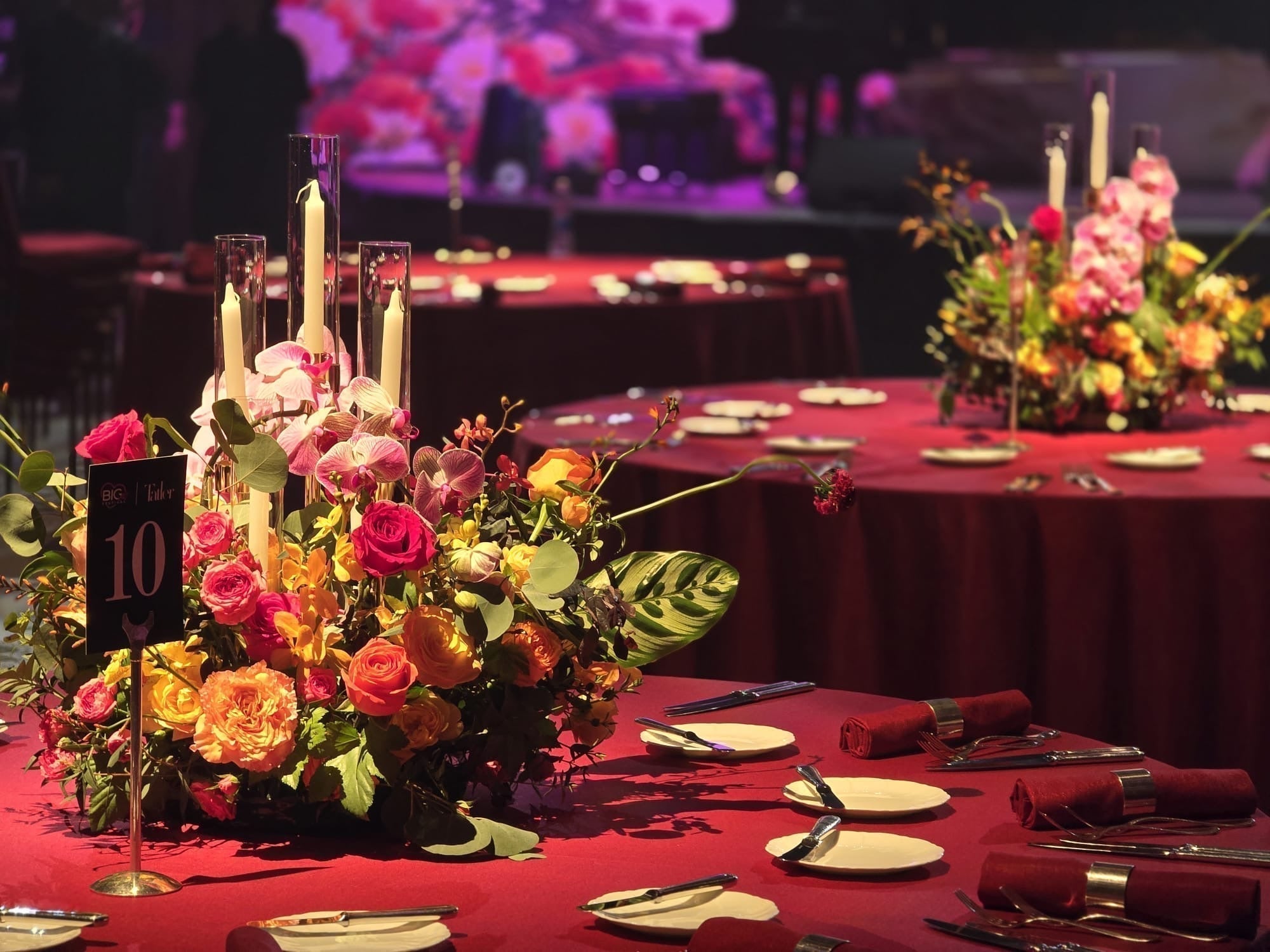 Gala Dinner 晚宴餐桌上的精緻花藝中心件設計，由 Lavish Florist 策劃。作品融合了典雅的粉色蝴蝶蘭、層次豐富的澄橙和黃色玫瑰，以及富有線條感的綠葉，呈現出溫暖華麗的色調。花藝設計巧妙融入真實燭台，讓燭光與鮮花交相輝映。