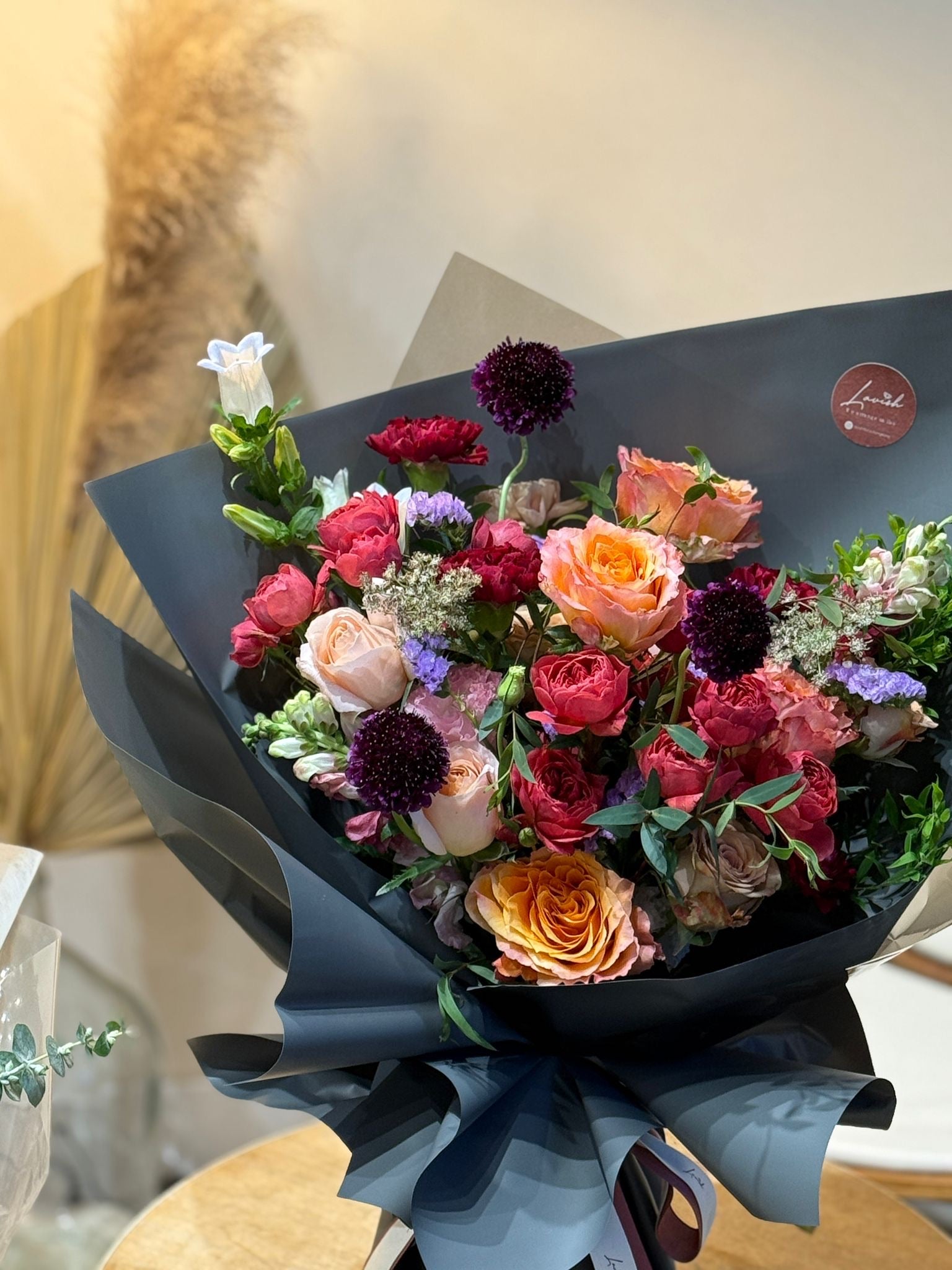 Bouquet of flowers wrapped in black paper with a visible brand logo. 精選男士花束近拍，展示了復古橙粉色玫瑰與深紫色松蟲草，包裹在高級霧面黑色包裝紙中。