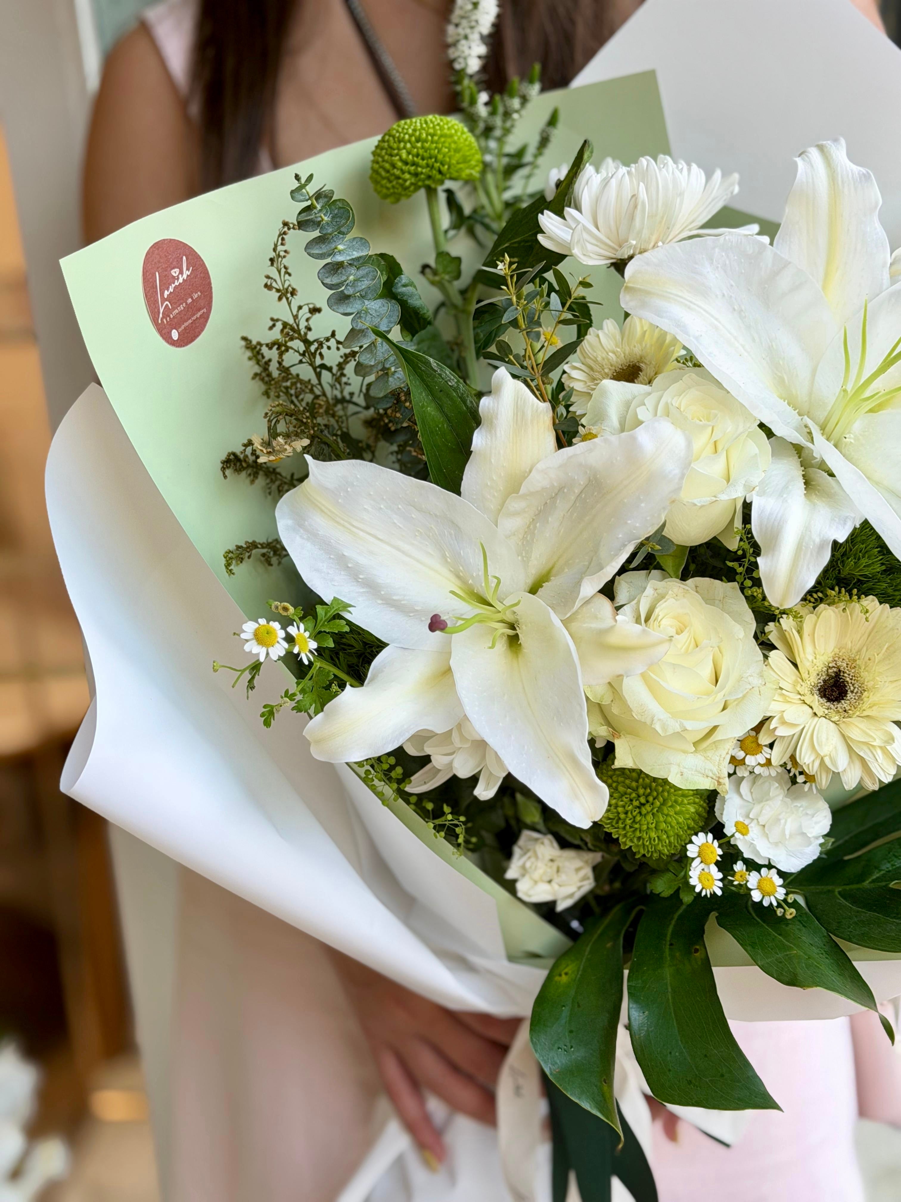 專業女花藝師手捧主題白綠色花束。這款花束由白百合、玫瑰、非洲菊和豐富的葉材手工綁紮而成。這是一份具有清新自然美感的精緻花藝作品，非常適合作為送給摯愛的禮物。