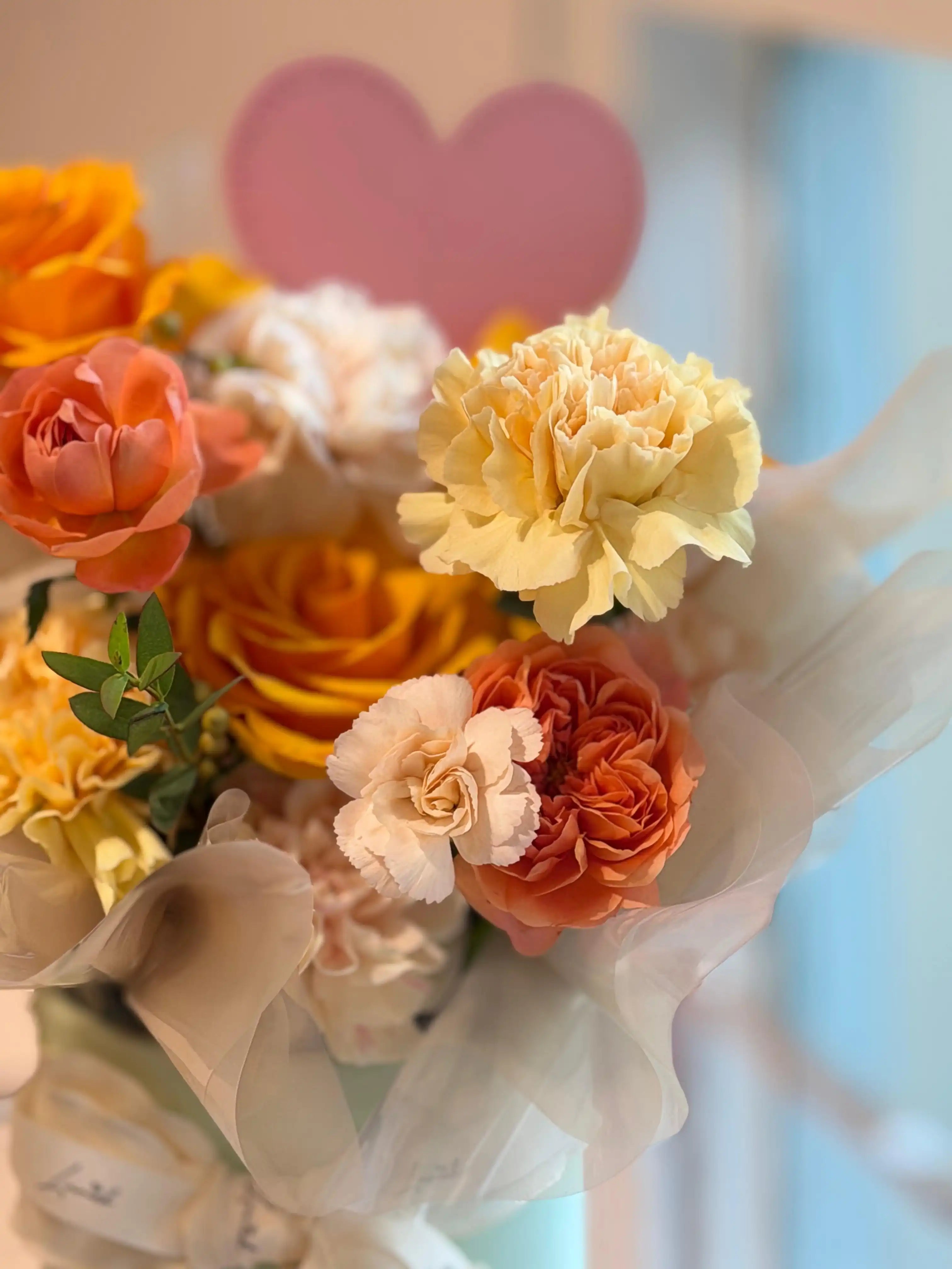 Luxury floral arrangement in a mint box with warm amber and coral roses, soft pink carnations, and a decorative heart pick.