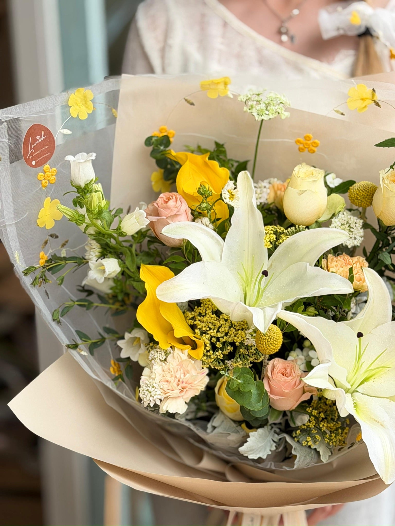 A deluxe cream-style bouquet called "Honeydew Lily" featuring large white lilies, yellow calla lilies, cream roses, and yellow dancing lady orchids in a translucent flower-patterned wrap.